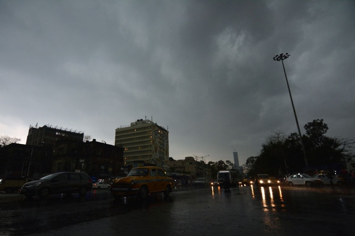 Dark cloudy sky over a Kolkata street with buildings, cars, yellow taxis, and wet roads reflecting headlights during rainy weather.