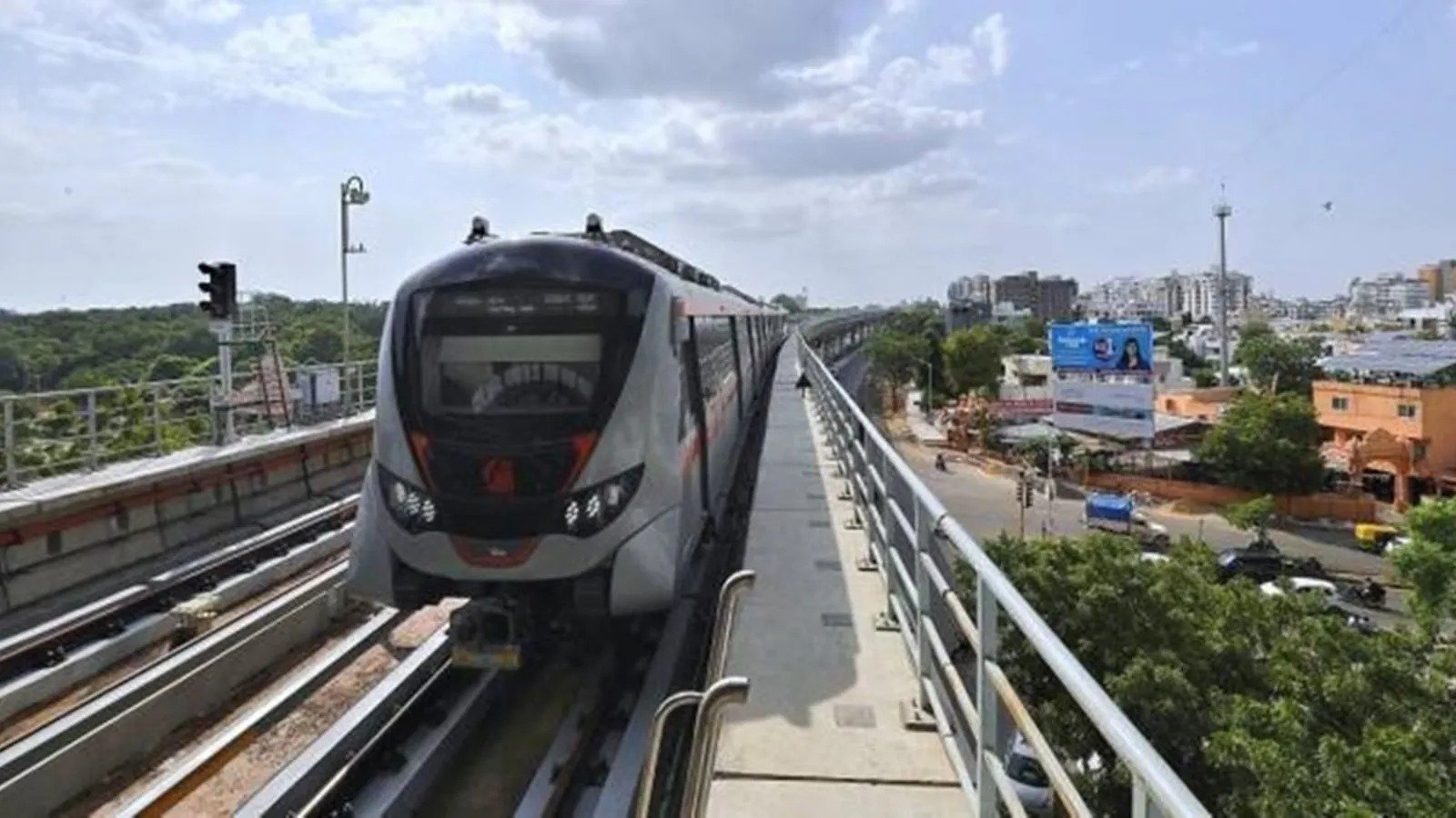 Metro train traveling on an elevated railway track above a city road, with buildings and traffic visible in the background.