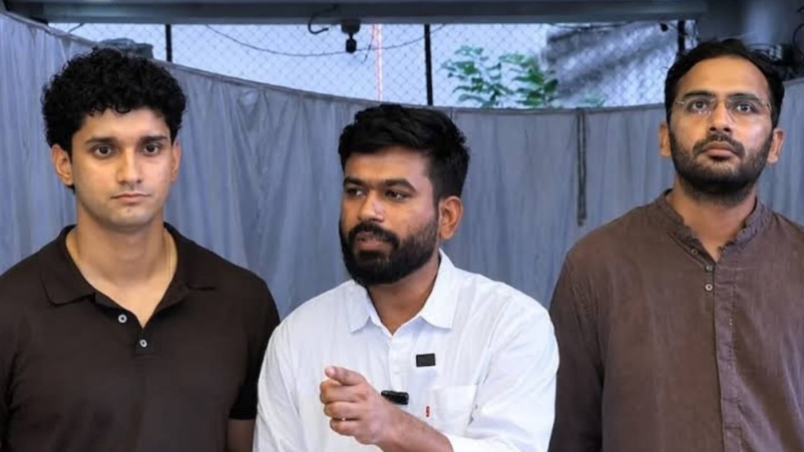 Three men standing side by side indoors, with the man in the center wearing a white shirt and holding a small object.
