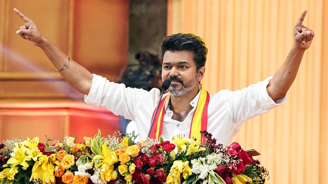 Tamil actor and TVK party leader Thalapathy Vijay addressing a public gathering, smiling and raising both hands amid a decorated stage with floral arrangements.