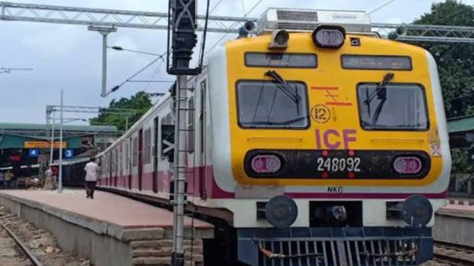 A yellow-and-white local passenger train standing on the railway tracks at a suburban station in India.