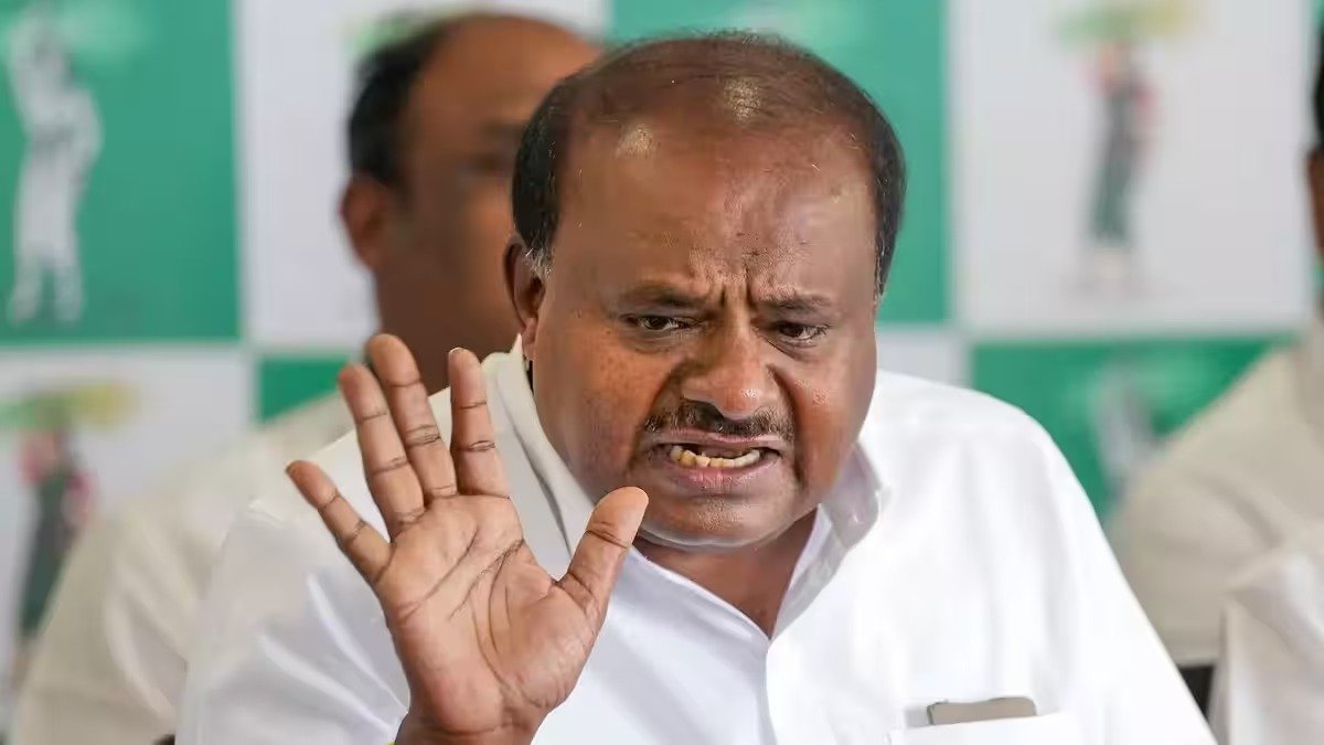 HD Kumaraswamy raising his hand during a public interaction, with a political party banner visible in the background.