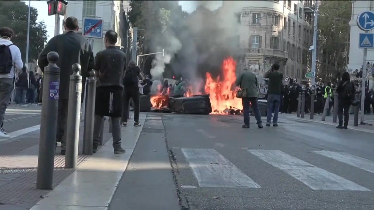 Street unrest in France with a fire burning on the road as people gather nearby amid ongoing protests.