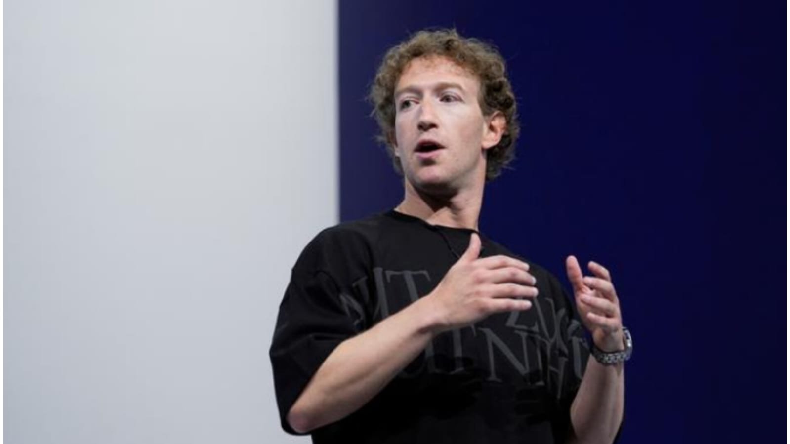 A man with curly brown hair wearing a black oversized T-shirt and a wristwatch speaks on stage, gesturing with both hands. He is looking slightly to the side with his mouth open mid-speech. The background features a large screen divided into light grey and dark navy blue sections.