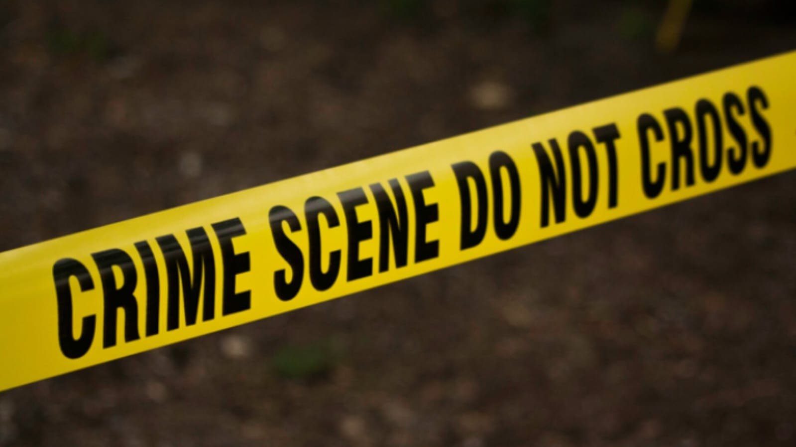 Close-up of a yellow crime scene tape with bold black text that reads "CRIME SCENE DO NOT CROSS" stretched across, with blurred ground in the background.