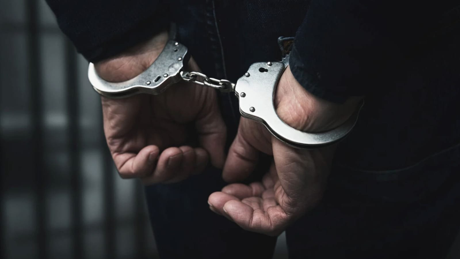 Close-up of a person's hands handcuffed behind their back, wearing silver metal handcuffs connected by a chain. The person is wearing a dark jacket, with blurred jail bars in the background.