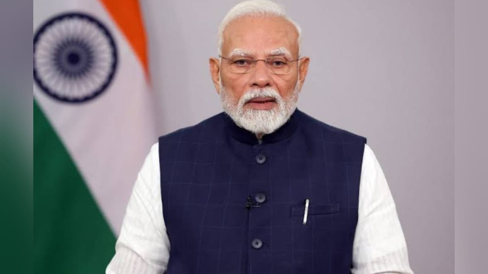 Prime Minister Narendra Modi speaking, wearing a white kurta and dark blue vest with a pen in pocket, Indian national flag visible in background.