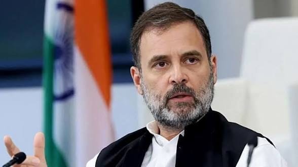 Congress leader Rahul Gandhi speaking at an event, wearing a white kurta and black vest, with the Indian national flag visible in the background.