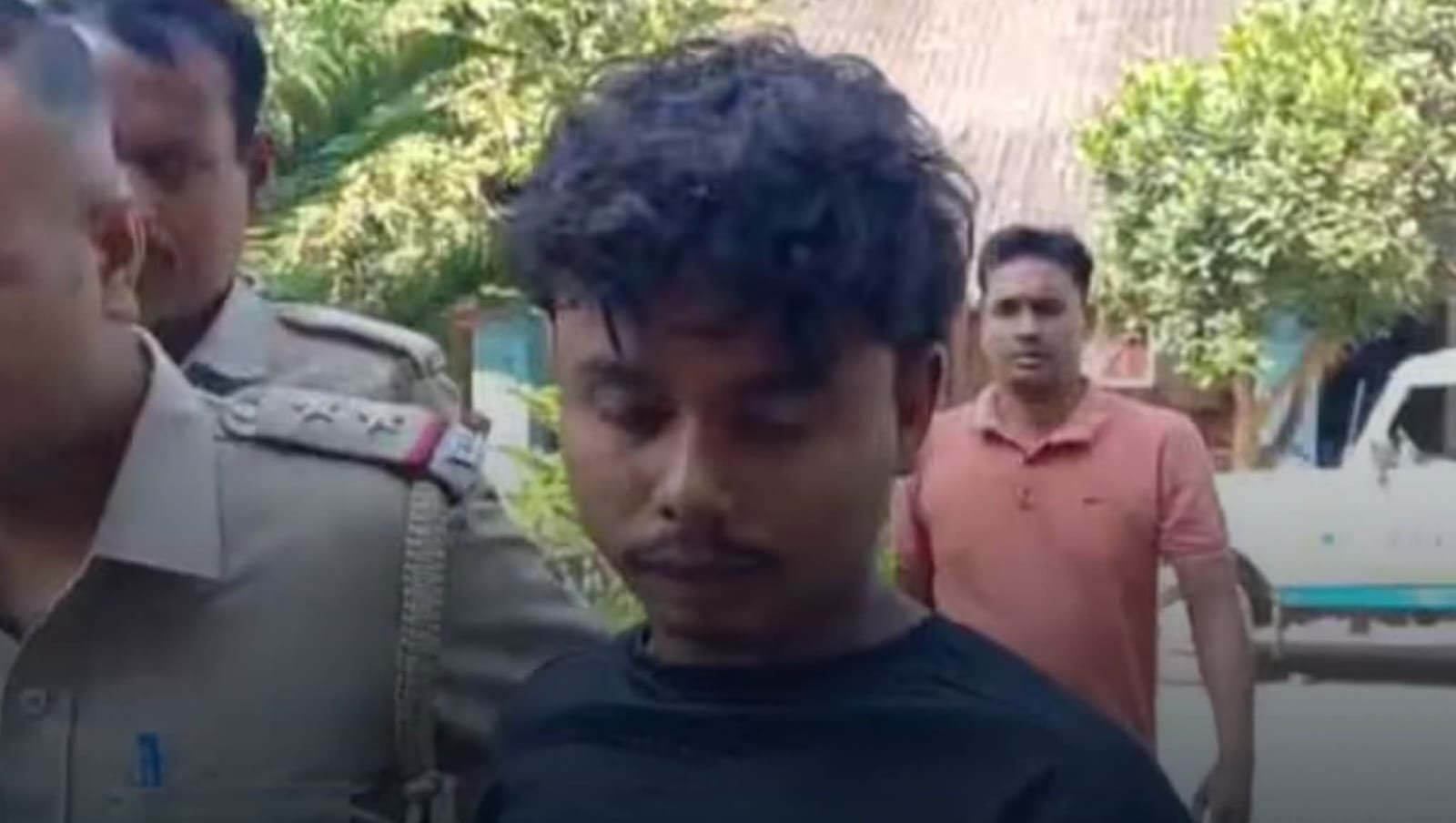 A young man in a black t-shirt being escorted by police officers in khaki uniform, outdoors with trees and a police vehicle visible in the background.
