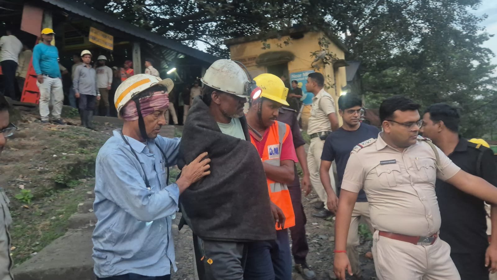 Rescue workers in helmets assisting a man wrapped in a blanket near a mine entrance, with police officers and other personnel in the background.
