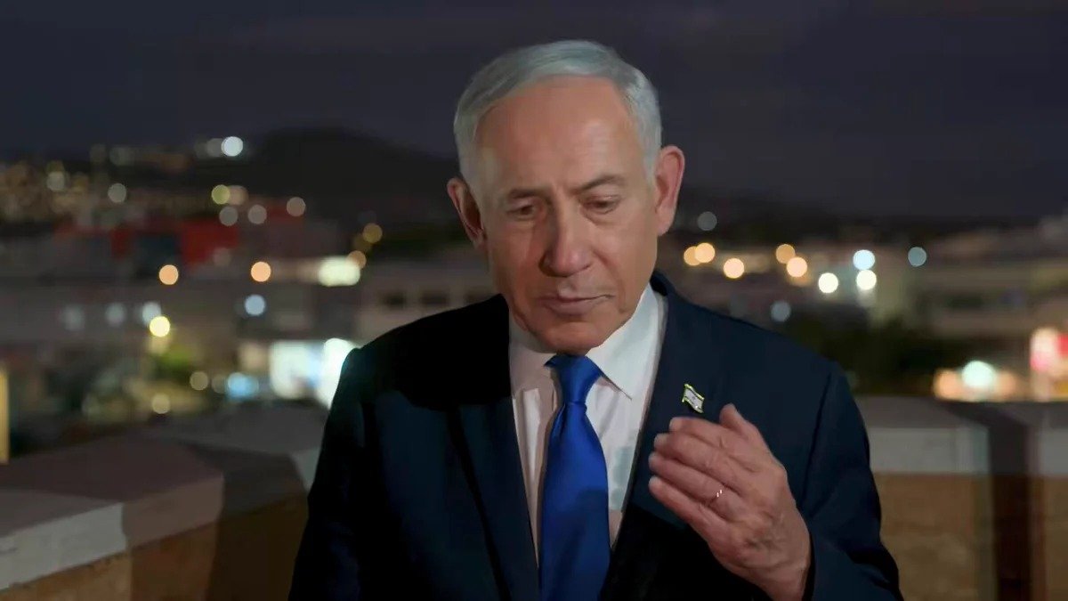 Israeli Prime Minister Benjamin Netanyahu, an older man with short white hair wearing a dark suit, white shirt and bright blue tie with a small Israeli flag pin on his lapel, standing outdoors at night on a balcony or rooftop with city lights blurred in the background, his left hand raised near his chest.