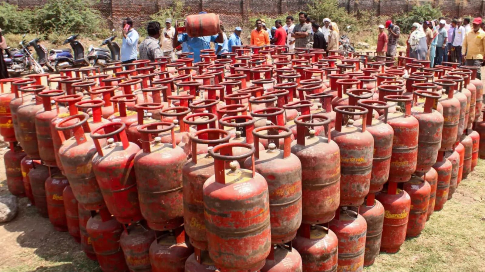 Large number of red LPG cooking gas cylinders stacked in rows outdoors on grassy ground, with a group of people standing in the background near a brick wall, one person carrying a cylinder on his shoulder.