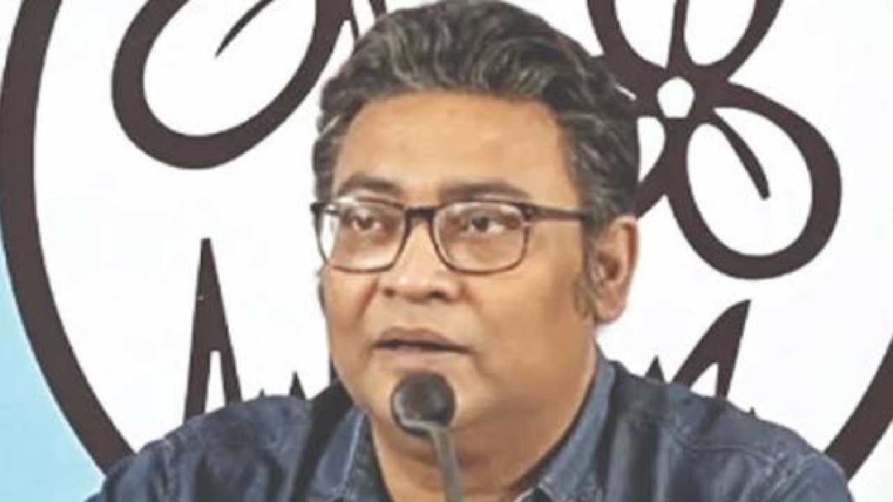 Close-up of a man with glasses and slightly graying hair, wearing a dark blue shirt, speaking into a microphone, with the black Trinamool Congress party symbol (two flowers with grass) visible on a white background behind him.