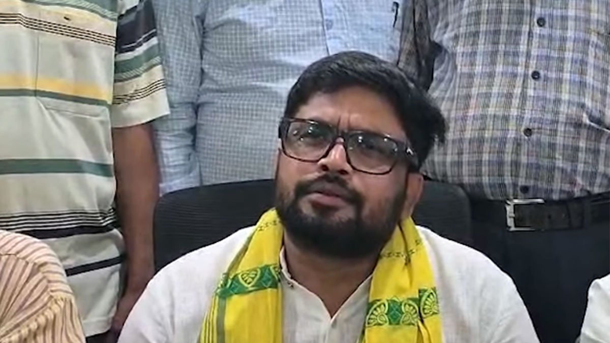 A man with black hair, beard and glasses wearing a white kurta and yellow scarf with green pattern sitting on a black chair. Three other people standing behind him out of focus.