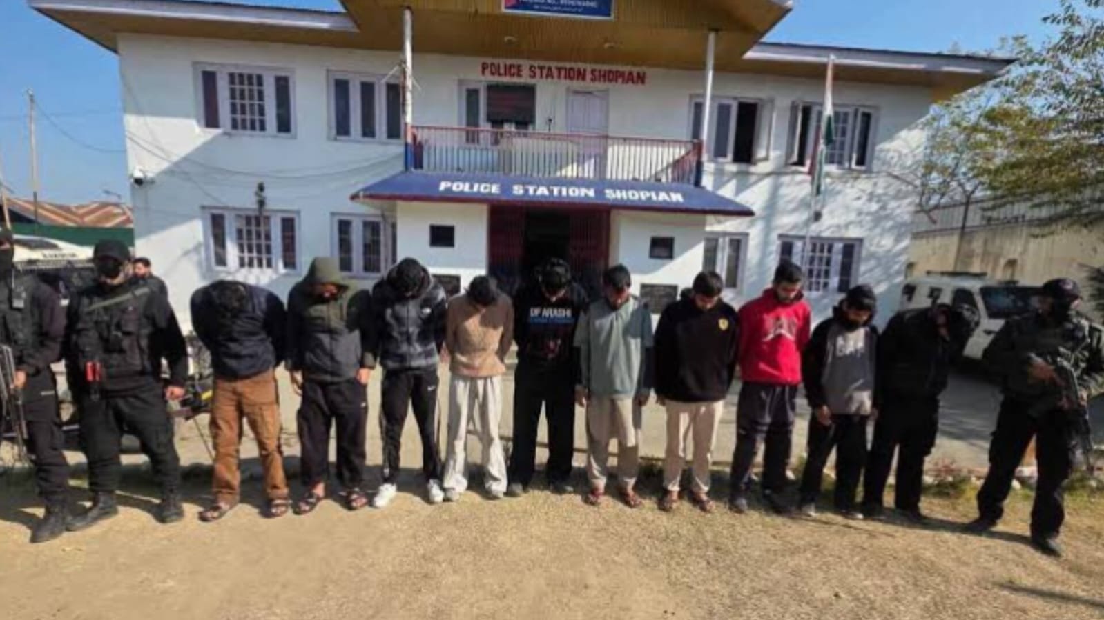 Ten arrested persons standing in front of Shopian Police Station in Jammu and Kashmir guarded by armed security personnel
