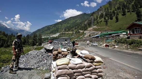 An Indian Army soldier stands guard beside sandbag bunkers on a mountain highway in Jammu and Kashmir, with green hills and a security checkpoint in the background.