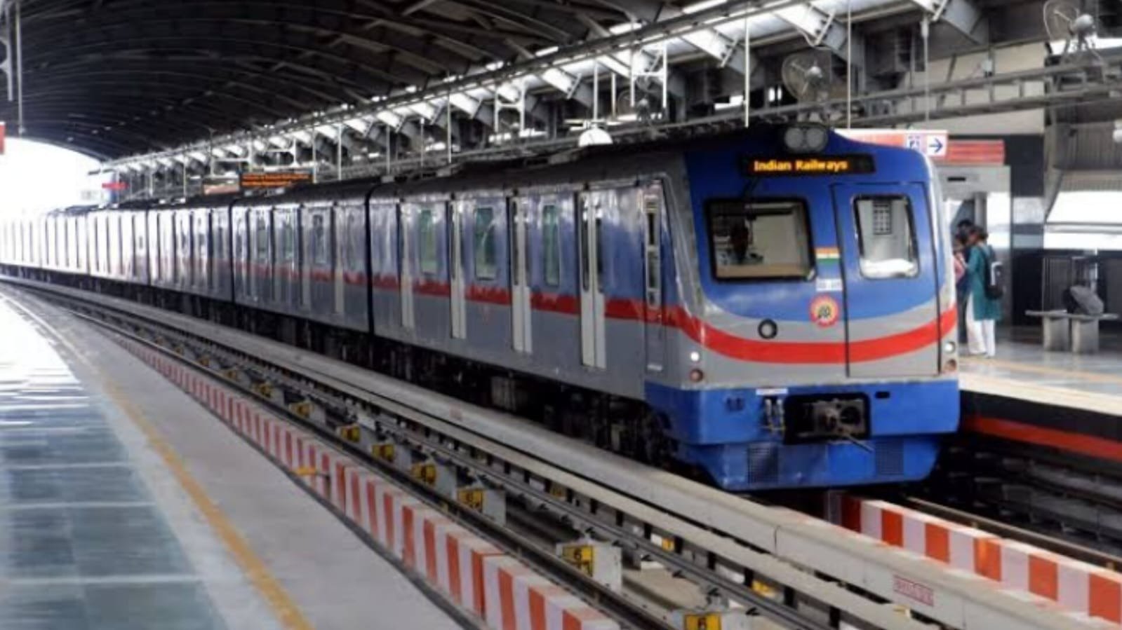 A blue, silver and red Kolkata Metro train arriving at an elevated station platform with passengers waiting on the platform.
