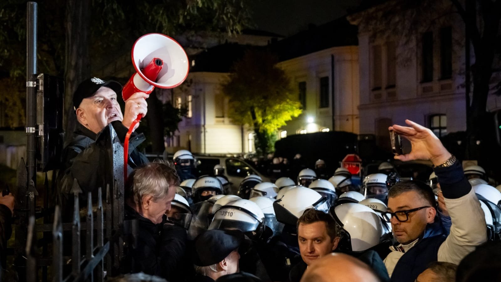 At night, a man in a black cap shouts through a red and white megaphone over a metal fence while facing a crowd of police officers in white riot helmets, with protesters gathered around recording the confrontation on phones.