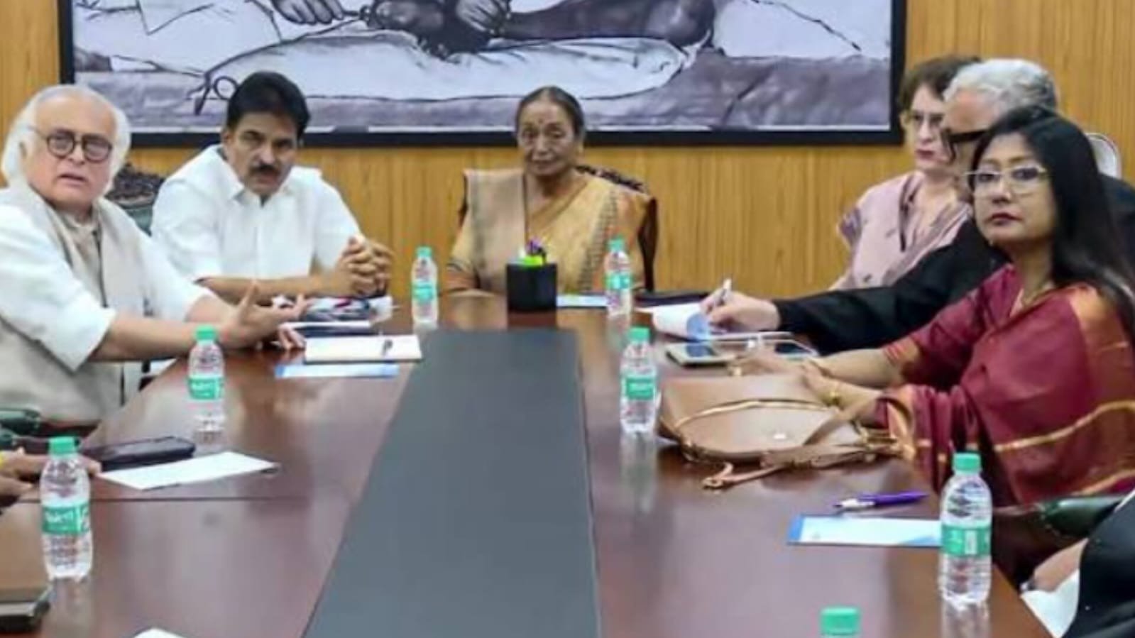 Five people, including Jairam Ramesh and KC Venugopal, sitting around a conference table in a meeting with water bottles and documents, with a framed picture of Mahatma Gandhi on the wall behind them.