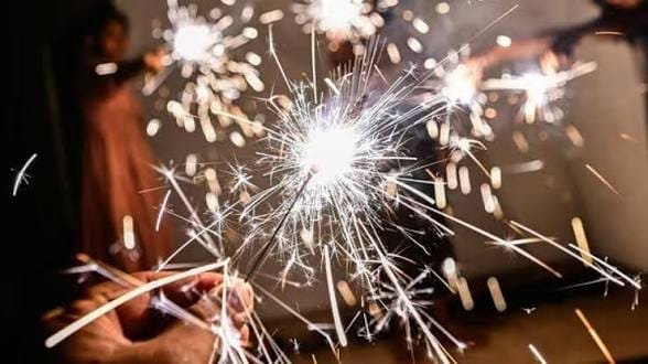 Close-up of hands holding brightly burning Diwali sparklers (phuljhuri) emitting white sparks, with blurred figures holding sparklers in the background at night.
